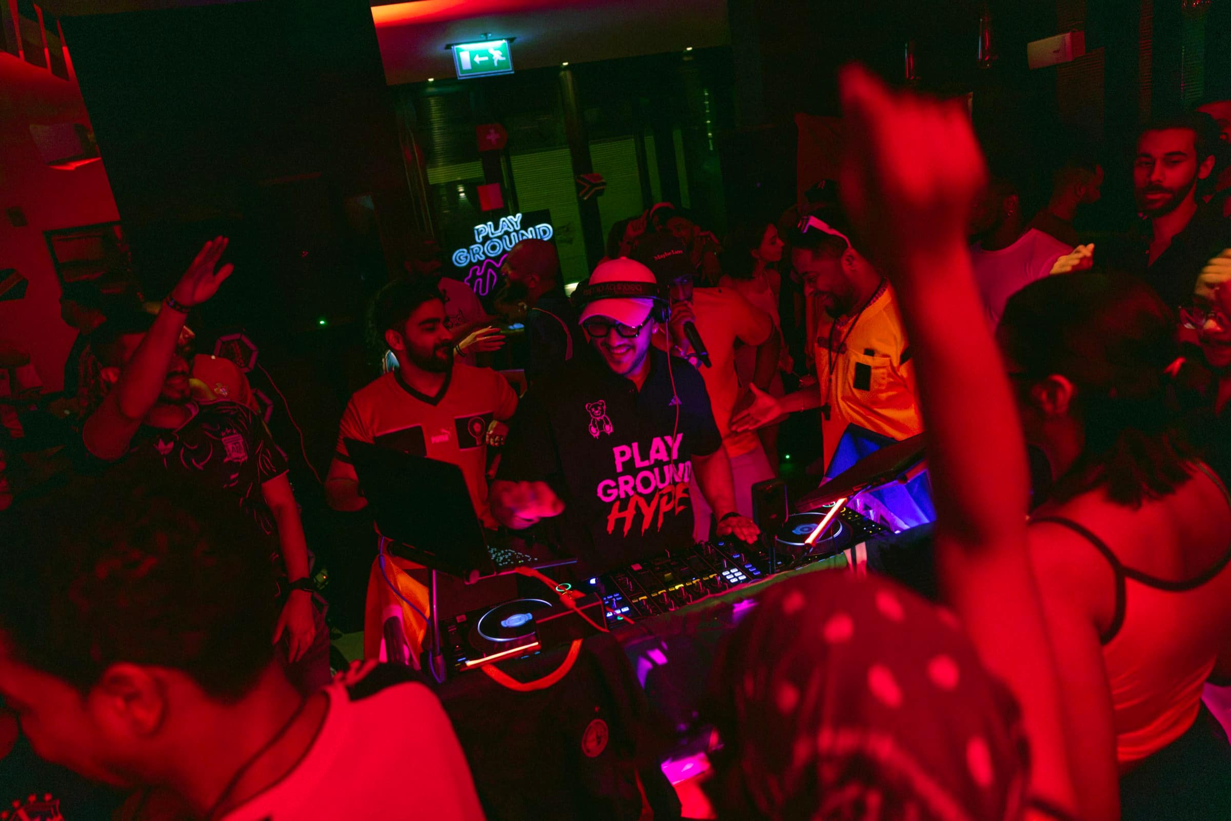 The floor at an OFFSIDE night — hands up around the decks, PLAYGROUND HYPE neon on the back wall.