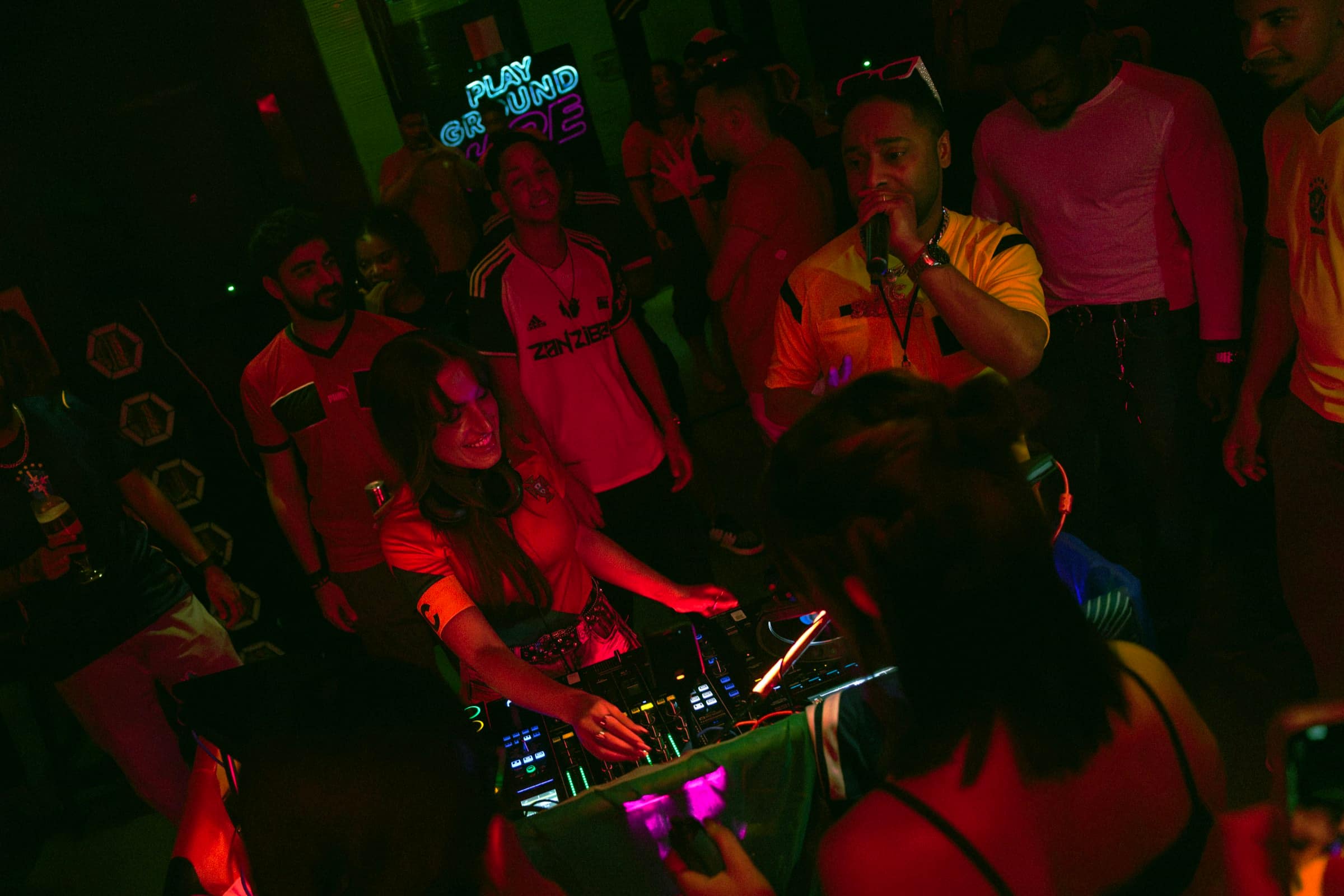A DJ mid-set surrounded on all sides by the crowd, jerseys and pink light.